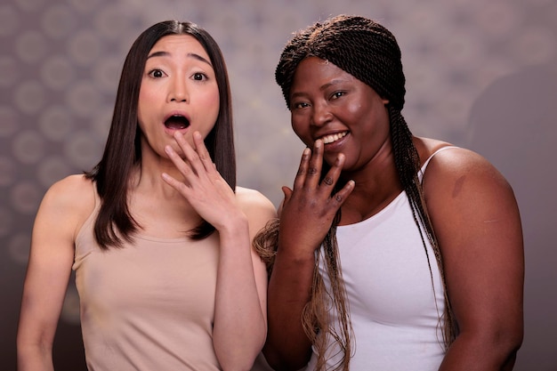 Photo two diverse surprised women laughing with hand over mouth, hearing funny joke. african american and asian beautiful girls giggling with shocked facial expression portrait