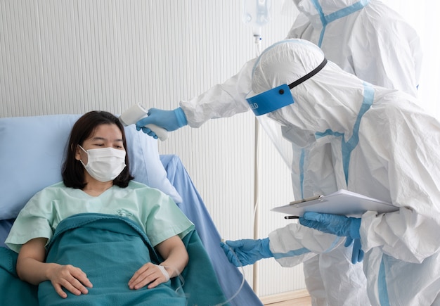 Two doctor wear PPE suit with face mask, measure body temperature of coronavirus infected patient with infrared thermometer gun in negative pressure quarantine room.