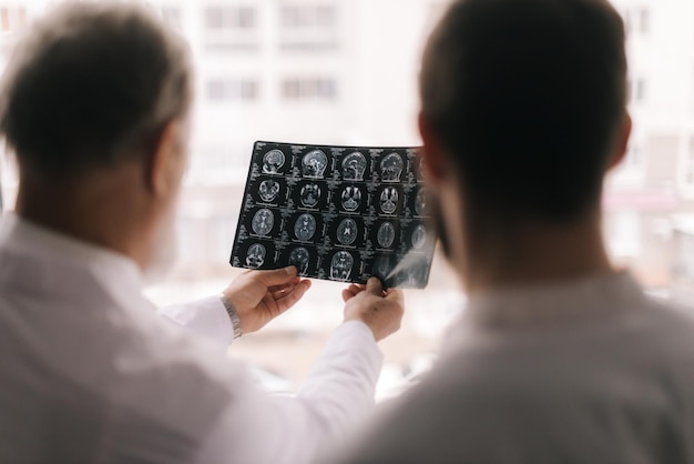 Two doctors discusses the results of an MRI scan of the patient's head in the consulting room against a large window. View from the doctors' backs, the focus on the X-ray. Concept of team medical work