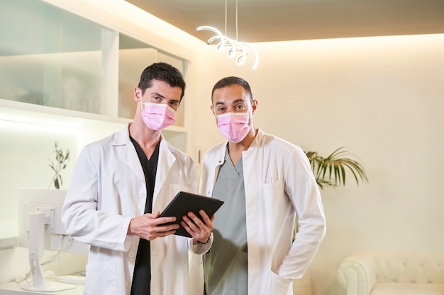 Two doctors in a mask looking at the camera with a tablet in hand. Gynecological, dental or aesthetic clinic. Medical concept.