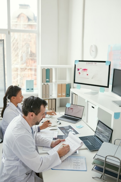 Two doctors sitting at the table and making notes in documents together during their work at office