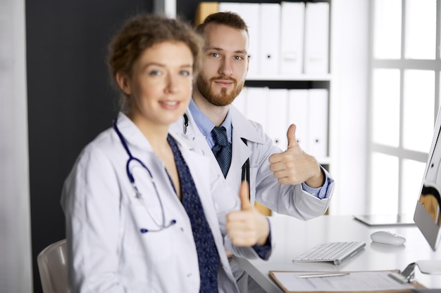 Два врача сидят с поднятыми большими пальцами в кабинете больницы. Медицинская помощь, противодействие вирусной инфекции и концепция медицины.