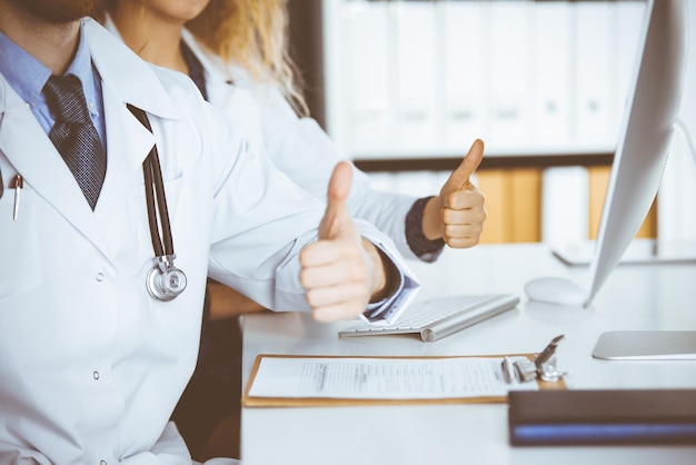 Two doctors sitting with thumbs up in hospital office. Medical help, countering viral infection and medicine concept.