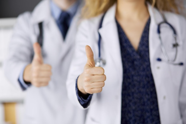 Two doctors standing with thumbs up in hospital office. Medical help, countering viral infection and medicine concept.