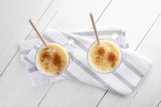 Two eggnog glasses with cinnamon and paper drinking straws on striped napkin on white wooden table