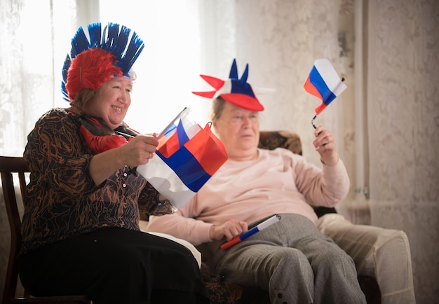 Two elderly enthusiastic women sitting in russian accessories and waving with russian flags