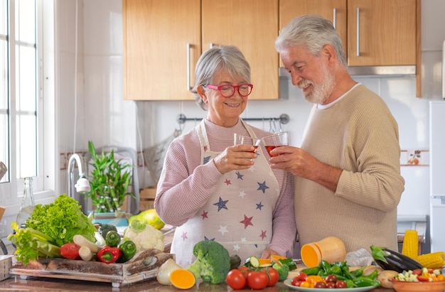 Due anziani preparano insieme le verdure in cucina, brindano con un bicchiere di vino rosso e sorridono. coppia senior allegra che si gode uno stile di vita vegetariano