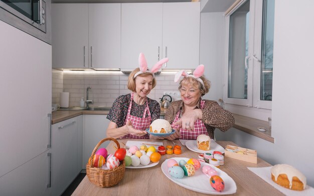 Two elderly women with rabbit ears on their heads are painting Easter eggs at home in the kitchen