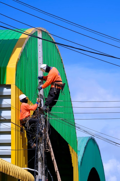 Two electricians installing electric cable lines on power pole near colorful warehouse building