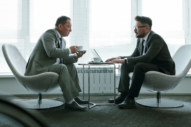 Two elegant businessmen in suits sitting in armchairs against large office window and discussing terms of contract or new strategies