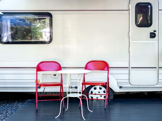 Two empty red iron chairs with white round table in front of the door and window of caravan car with nobody Relaxation camping and sleep in the motorhome trailer Family vacation travel concept