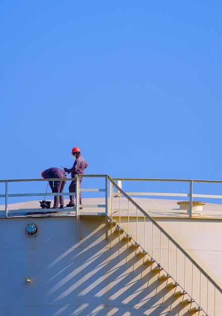 Two engineers checking oil quality on top of oil fuel storage tank against blue sky background