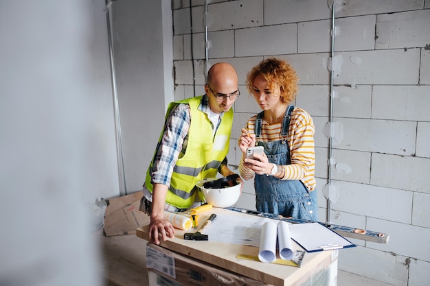 Foto due ingegneri che guardano lo schermo del telefono, sporgendosi dal tavolo con il progetto. accanto a un muro di mattoni all'interno di un edificio in costruzione.