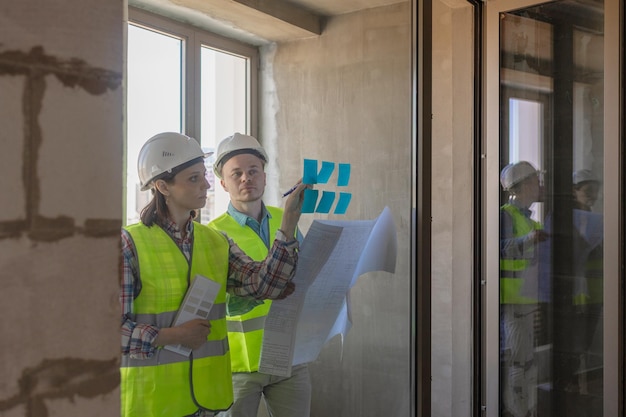 Two engineers in protective vests and white helmets glue stickers on the window and discuss the construction plan of the facility or the scheme