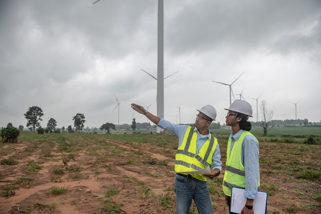 Two engineers working and holding the report at wind turbine farm Power Generator Station on mountainThailand people