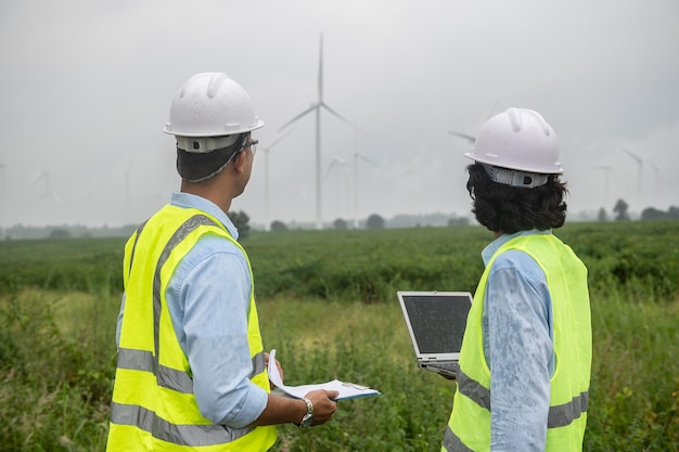 Two engineers working and holding the report at wind turbine farm Power Generator Station on mountainThailand people