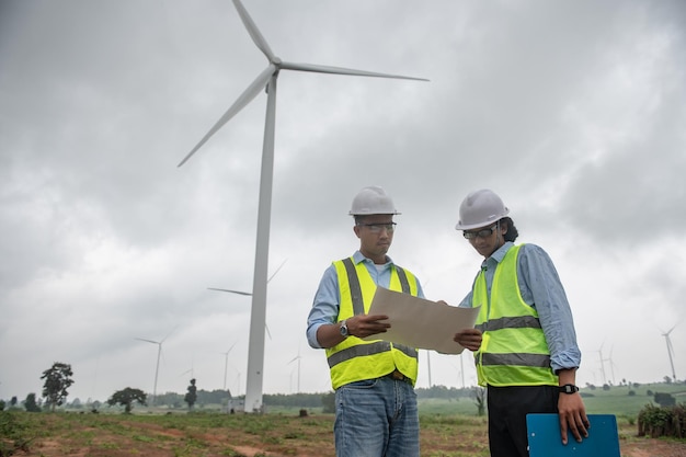 Two engineers working and holding the report at wind turbine farm Power Generator Station on mountainThailand people