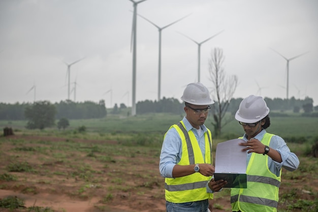 Two engineers working and holding the report at wind turbine farm Power Generator Station on mountainThailand people