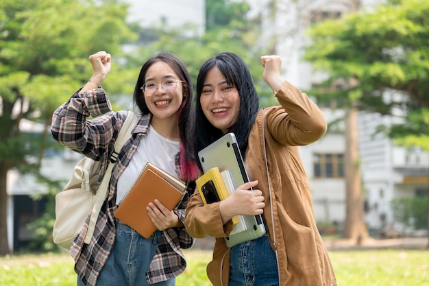 사진 두 명의 흥분된 아시아 여성 학생들이 야외에 서서 축하하기 위해 주먹을 들어 올리고 있습니다.