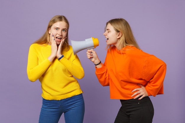 Две возбужденные веселые молодые блондинки сестры-близнецы в ярких красочных одеждах кричат на мегафон, изолированном на пастельно-фиолетовой синей стене. Концепция семейного образа жизни людей. .