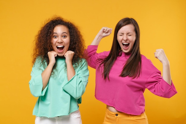 Two excited women friends european african american girls in pink green clothes posing isolated on yellow orange background. People lifestyle concept. Mock up copy space. Clenching fists like winner.