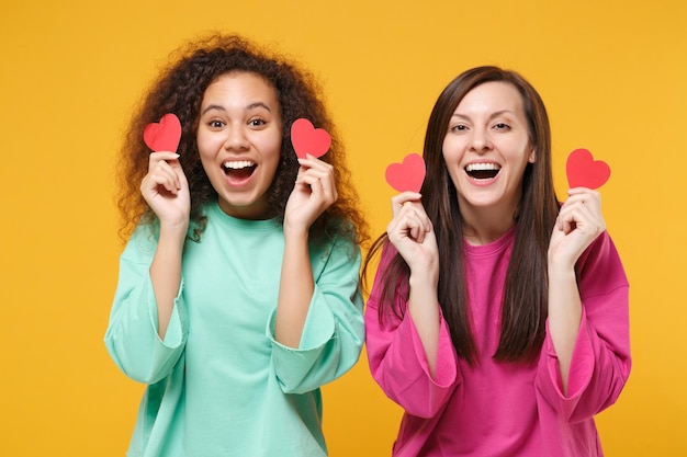 Photo two excited women friends european and african american girls in pink green clothes posing isolated on yellow wall background. people lifestyle concept. mock up copy space. holding little red hearts.