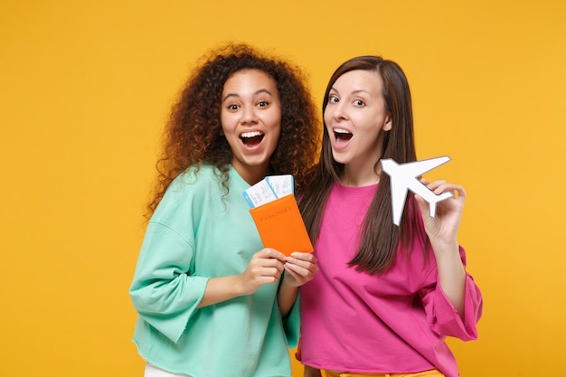 Two excited women friends european african girls in pink green clothes posing isolated on yellow background. People lifestyle concept. Mock up copy space. Hold passport boarding pass ticket, airplane.