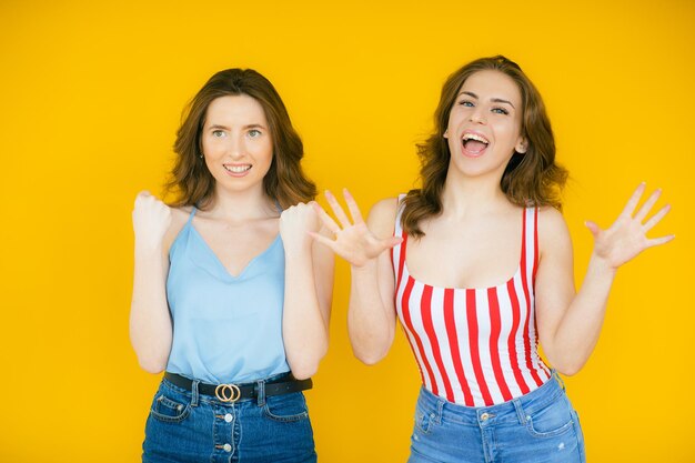 Two excited young girls dressed in summer clothes whispering secrets isolated over yellow