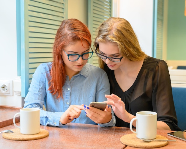 Two excited young girls using mobile phones sitting in a cafe and pointing finger Redhaired woman in glasses shows her friend funny photos on a smartphone girlfriend blonde They drink coffee