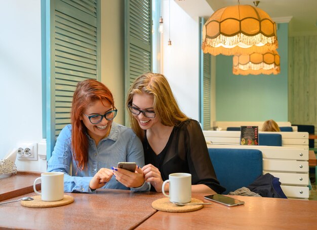 Two excited young girls using mobile phones sitting in a cafe and pointing finger Redhaired woman in glasses shows her friend funny photos on a smartphone girlfriend blonde They drink coffee