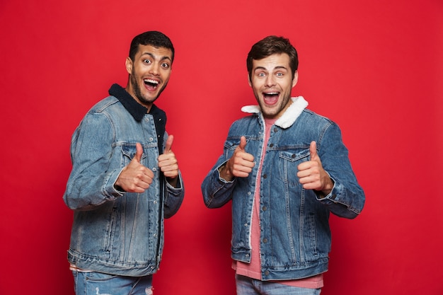 Two excited young men friends wearing denim jackets standing isolated over red wall, giving thumbs up