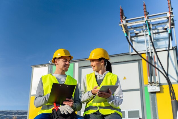 Two experienced workers woman and man in hard hats and signal vests look at tablet and check the operation of solar panels