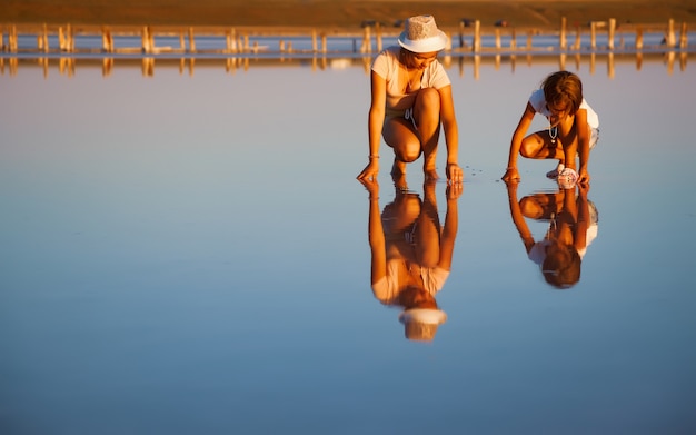 아름다운 투명한 소금 호수에 특이한 의상을 입은 두 명의 환상적으로 아름다운 소녀가 반짝이는 표면에서 무언가를 찾고 있습니다.