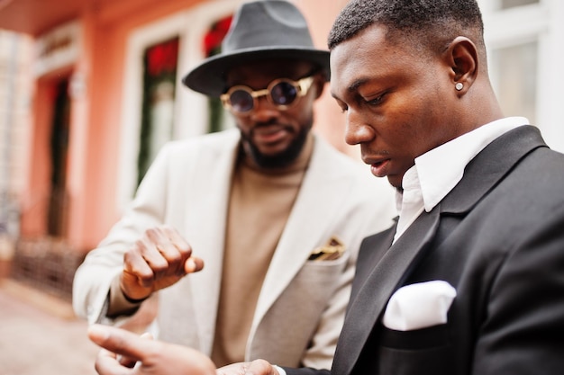 Two fashion black men stand near business car and look at cell phone Fashionable portrait of african american male models Wear suit coat and hat