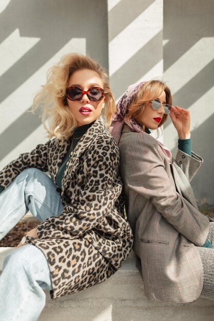 Two fashion glamorous young women girlfriends in trendy autumn clothes with sunglasses leopard blazer jeans and suit sits on the street in sunlight