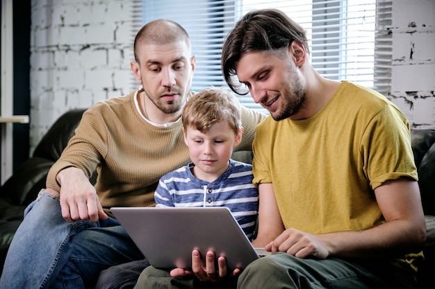 Two fathers and their preteen son spending time together at home and watching movie or playing game on laptop