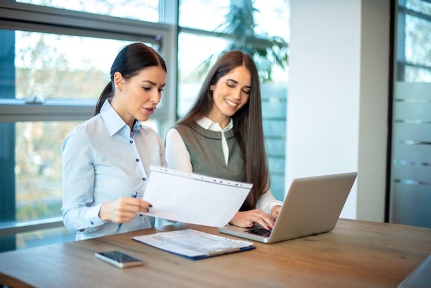 Two female business coworkers discussing a paper document and using laptop at office