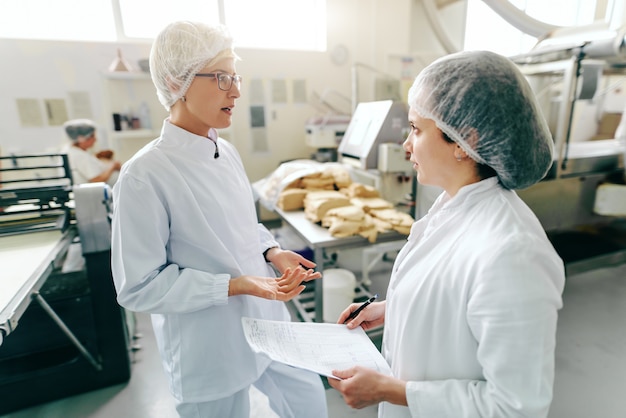 Due impiegati caucasici femminili che parlano del lavoro mentre stando nella fabbrica dell'alimento. uno di loro in possesso di documenti. nelle macchine in background.