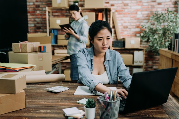 Две коллеги-женщины работают в современном офисе склада. сосредоточенная молодая азиатская девушка, печатающая на клавиатуре портативного компьютера, управляет своим интернет-магазином, малым бизнесом, отвечая на сообщение клиента в Интернете