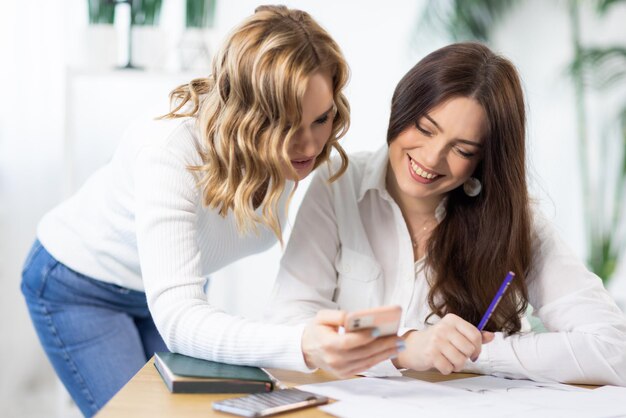 Two female employees are discussing the details of project using information from Internet on phone near table in office