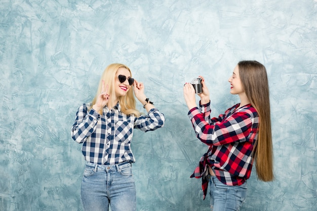 Due amiche in camicie a scacchi e jeans che fotografano con una fotocamera retrò sullo sfondo del muro dipinto di blu