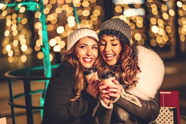 Photo two female friends enjoy the night out hugging and laughing with cups of coffee in their hands.