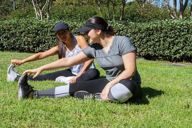 Two female friends in the park stretching their legs to start their workout while having fun