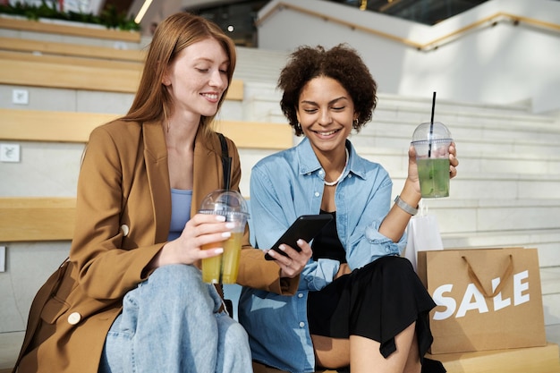 Two female friends sitting on bench at shopping mall chatting checking information on smartphone