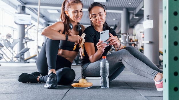 Two female friends in sportive clothes is in the gym earing fruits and using phone.