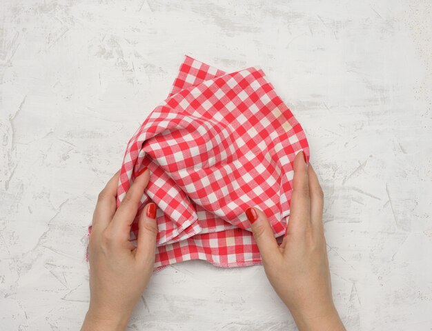 Two female hands hold a crumpled white-red checkered textile kitchen napkin on a white table, top view