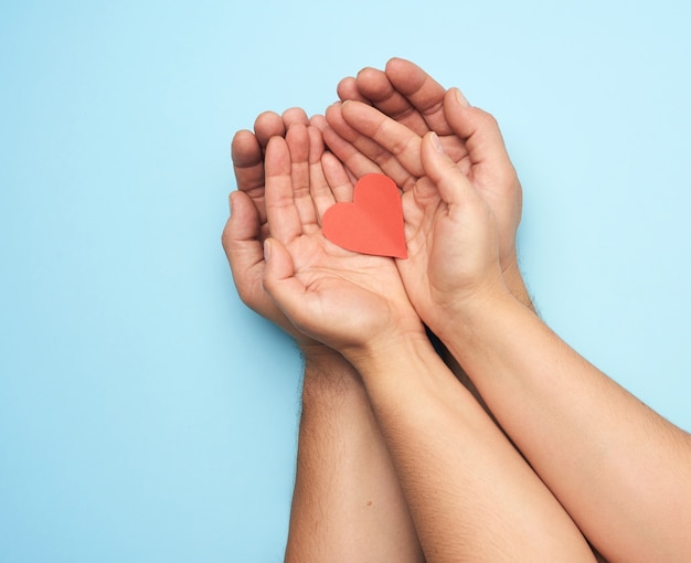 Photo two female hands lie in male palms and hold a red paper heart, top view. concept of kindness, love and donation