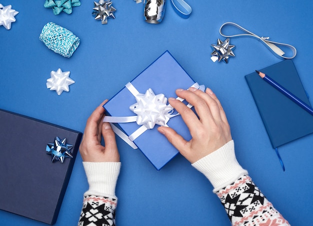 Two female hands in a sweater hold a square blue box with a bow