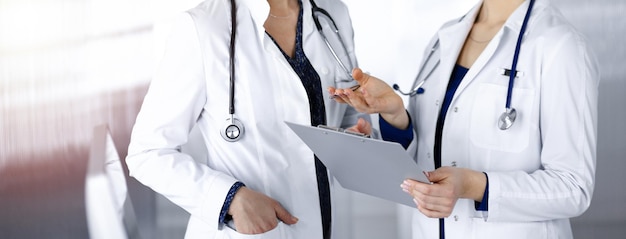 Two female physicians are discussing their patient's medical tests, while standing in a sunny clinic office. Doctors use a clipboard at work. Teamwork in medicine.