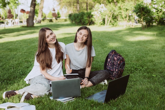 2人の女子学生が本とノートパソコンを持って芝生の公園に座って勉強し、試験の準備をしています。遠隔教育。ソフトセレクティブフォーカス。